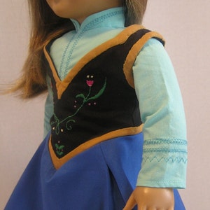 May include: A doll wearing a blue shirt with a black vest with brown trim and floral embroidery. The doll is also wearing a blue skirt.