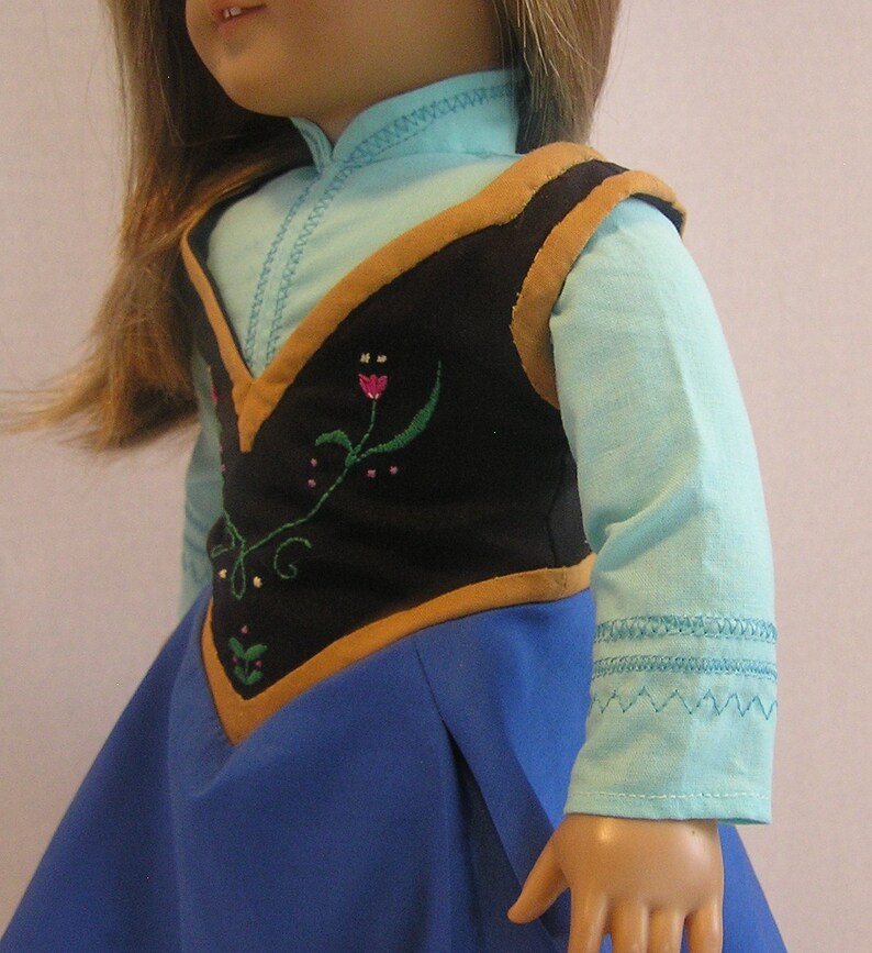May include: A doll wearing a blue shirt with a black vest with brown trim and floral embroidery. The doll is also wearing a blue skirt.