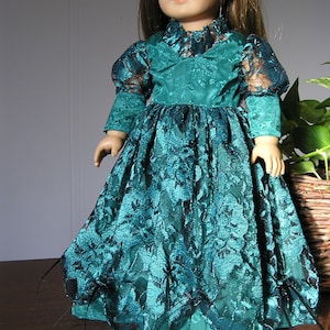 Puede incluir: Una mu&ntilde;eca con un vestido de encaje verde azulado con ribetes negros. El vestido tiene un corpi&ntilde;o ajustado y una falda completa. La mu&ntilde;eca tiene el pelo rubio y los ojos azules.