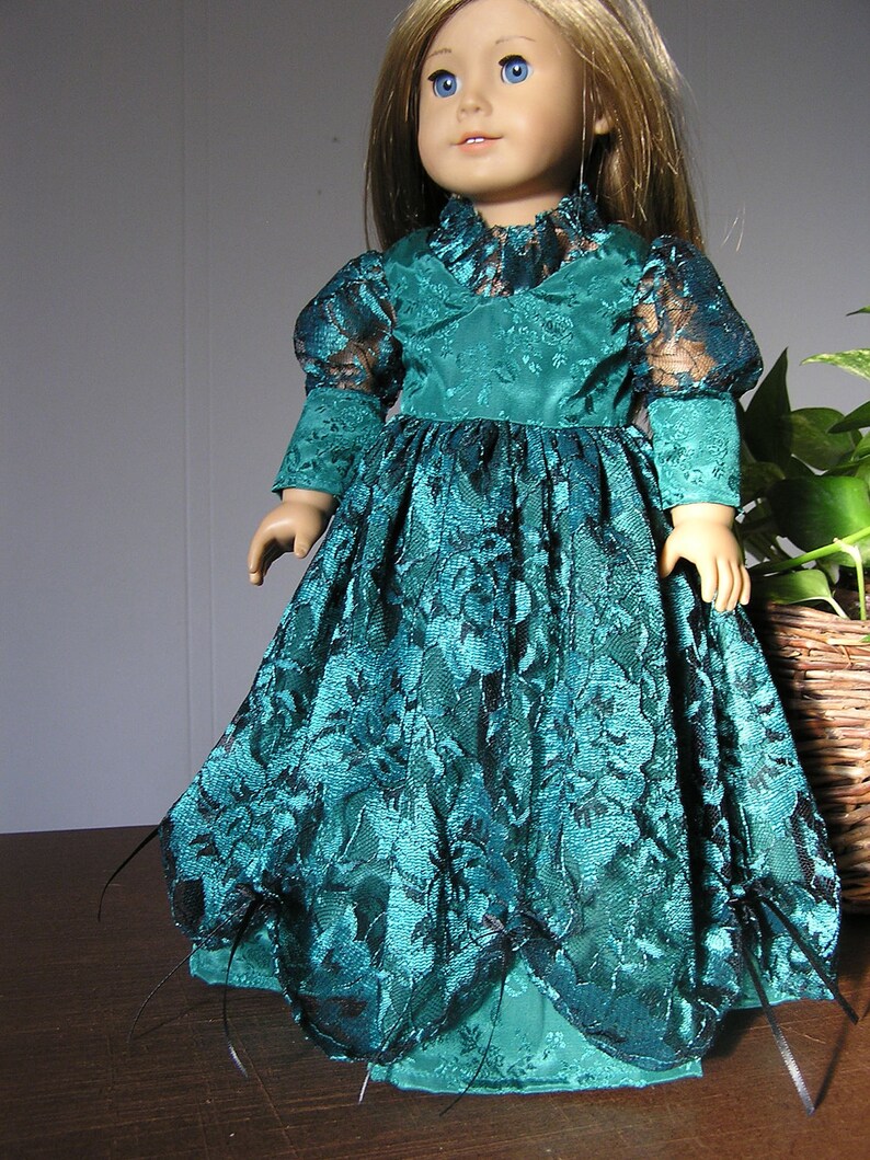 Puede incluir: Una mu&ntilde;eca con un vestido de encaje verde azulado con ribetes negros. El vestido tiene un corpi&ntilde;o ajustado y una falda completa. La mu&ntilde;eca tiene el pelo rubio y los ojos azules.