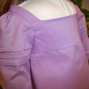 May include: A lavender dress with long sleeves and a gathered detail at the shoulders. The dress has a square neckline and a simple, elegant design.