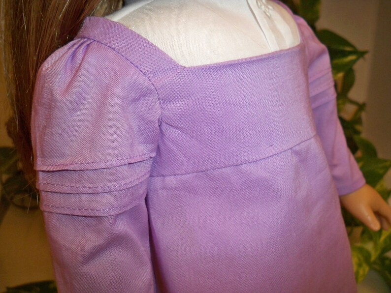 May include: A lavender dress with long sleeves and a gathered detail at the shoulders. The dress has a square neckline and a simple, elegant design.