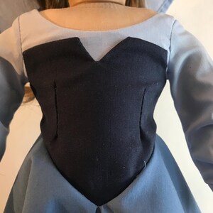 K&ouml;nnte beinhalten: Ein Puppenkleid mit einem marineblauen Oberteil und einem hellblauen Rock und &Auml;rmeln. Das Oberteil hat einen V-Ausschnitt und ist tailliert. Die &Auml;rmel sind lang und das Kleid scheint handgefertigt zu sein.