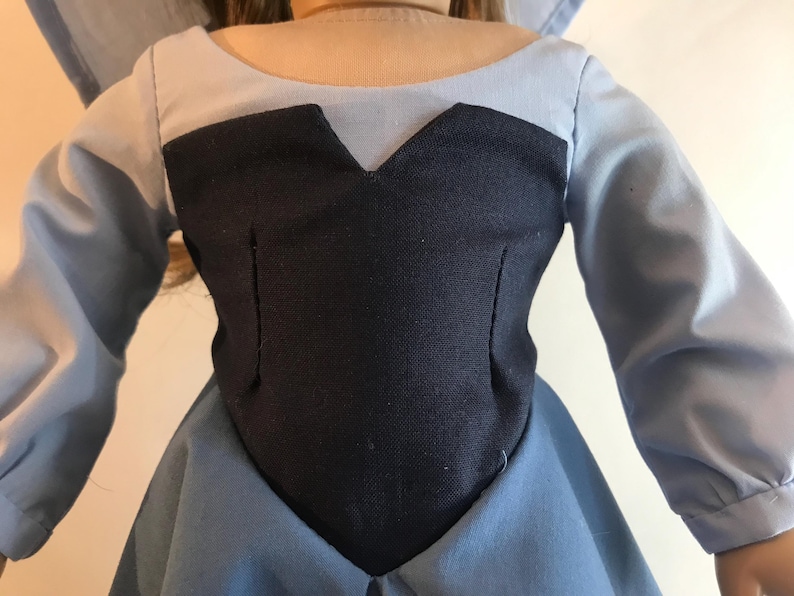 K&ouml;nnte beinhalten: Ein Puppenkleid mit einem marineblauen Oberteil und einem hellblauen Rock und &Auml;rmeln. Das Oberteil hat einen V-Ausschnitt und ist tailliert. Die &Auml;rmel sind lang und das Kleid scheint handgefertigt zu sein.