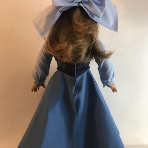 K&ouml;nnte beinhalten: Eine Puppe in einem hellblauen Kleid mit langen &Auml;rmeln und einer gro&szlig;en passenden Schleife. Die Puppe hat braunes Haar, und das Kleid hat ein dunkelblaues Oberteil. Die Puppe wird von hinten betrachtet.