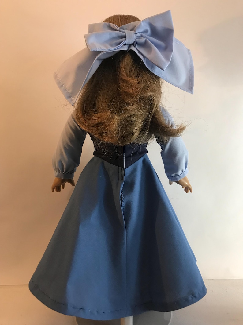 K&ouml;nnte beinhalten: Eine Puppe in einem hellblauen Kleid mit langen &Auml;rmeln und einer gro&szlig;en passenden Schleife. Die Puppe hat braunes Haar, und das Kleid hat ein dunkelblaues Oberteil. Die Puppe wird von hinten betrachtet.