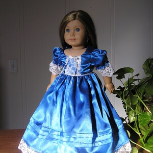 Puede incluir: Un vestido de mu&ntilde;eca de sat&eacute;n azul con ribete de encaje blanco. El vestido tiene un corpi&ntilde;o ajustado y una falda completa. La mu&ntilde;eca lleva un cuello de encaje blanco.