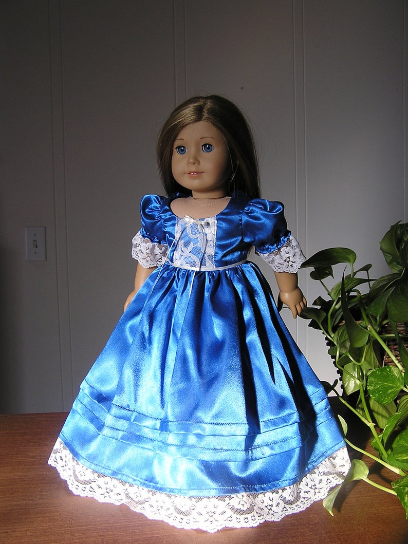 Puede incluir: Un vestido de mu&ntilde;eca de sat&eacute;n azul con ribete de encaje blanco. El vestido tiene un corpi&ntilde;o ajustado y una falda completa. La mu&ntilde;eca lleva un cuello de encaje blanco.