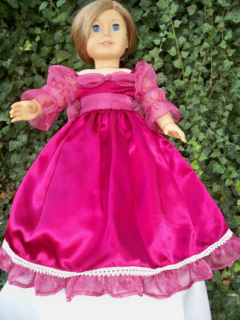 Puede incluir: Un vestido de mu&ntilde;eca de sat&eacute;n rosa con ribete de encaje blanco y mangas transparentes. El vestido tiene un corpi&ntilde;o ajustado y una falda completa.