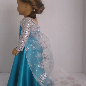 Puede incluir: Un vestido azul turquesa con un corpi&ntilde;o de lentejuelas plateadas y una capa blanca transparente con dise&ntilde;os de copos de nieve. El vestido es para una mu&ntilde;eca.