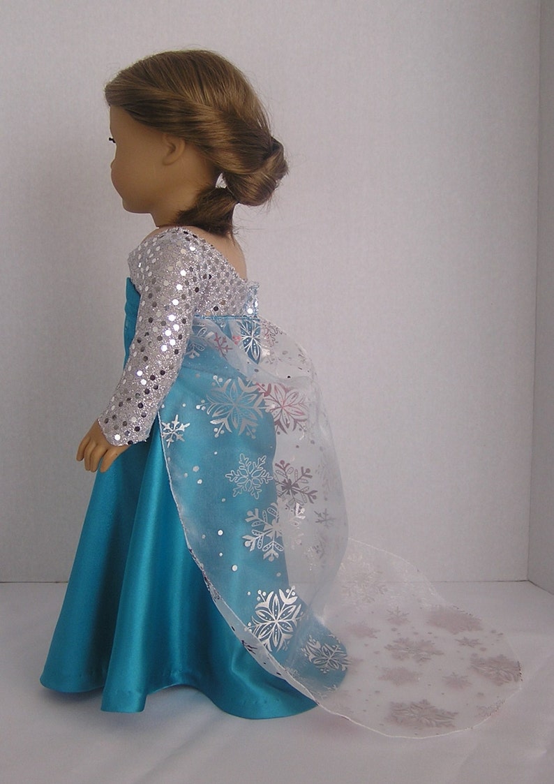 Puede incluir: Un vestido azul turquesa con un corpi&ntilde;o de lentejuelas plateadas y una capa blanca transparente con dise&ntilde;os de copos de nieve. El vestido es para una mu&ntilde;eca.