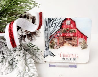 May include: A festive square decoration with a red barn scene, snow, and the words "Christmas on the Farm." The image also shows a tree and a pine cone with red and white ribbon, evoking a winter theme.