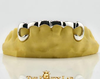 Op de afbeelding: Een zilverkleurige, op maat gemaakte grillz-set, gepresenteerd op een gele tandheelkundige mal. De grillz hebben een gepolijste afwerking en een gebogen ontwerp. Het logo "THE KWITY LAB CUSTOM GRILLZ FINE" is onderaan zichtbaar.