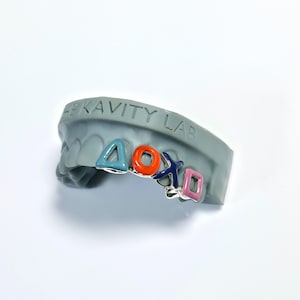 May include: A silver grill with colorful enamel designs in the shape of video game controller buttons. The grill is displayed on a gray dental mold. The text "THE KAVITY LAB" is visible on the mold.