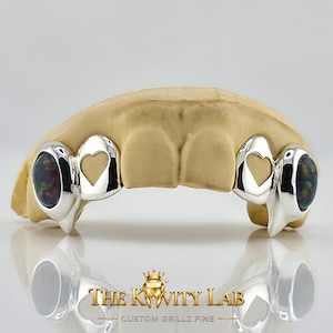 May include: A silver dental grill set featuring two outer teeth with embedded dark iridescent opal stones and two inner teeth with heart-shaped cutouts. The grill is displayed on a beige dental mold, with the text THE KWITY LAB CUSTOM GRILLZ FINE.