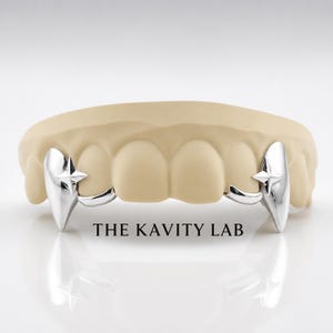 May include: A silver-colored grillz set featuring two pointed fang-like caps with four-pointed star engravings on the canine teeth, fitted onto a beige dental mold. The text THE KAVITY LAB is printed in black below the dental appliance.