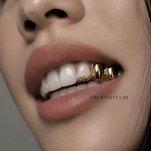 May include: A gold tooth grill featuring a smooth, polished finish on two teeth and a textured, ornate gold accent piece attached to the adjacent tooth. The text THE KAVITY LAB is visible in black lettering near the lower lip area of the mouth.