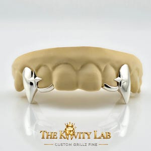 May include: Silver-colored custom grillz with a star design. The grillz are displayed on a beige dental mold. The text "THE KWITY LAB" and "CUSTOM GRILLZ FINE" are visible at the bottom of the image.