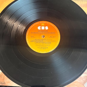 May include: A black vinyl record with a yellow and orange label featuring the text "KALAAKAAR" and "AN ORIGINAL SOUNDTRACK RECORDING." The record is on a patterned surface. The label includes song titles and credits.