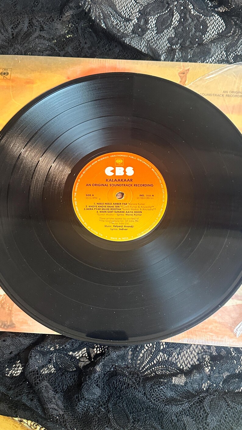 May include: A black vinyl record with a yellow and orange label featuring the text "KALAAKAAR" and "AN ORIGINAL SOUNDTRACK RECORDING." The record is on a patterned surface. The label includes song titles and credits.
