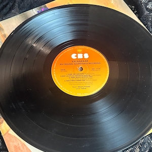 May include: A vintage vinyl record from the soundtrack of "Kalaakaar" by CBS. The black record is centered on a colorful label with text. The record is partially visible, with the album cover and a black lace fabric in the background.