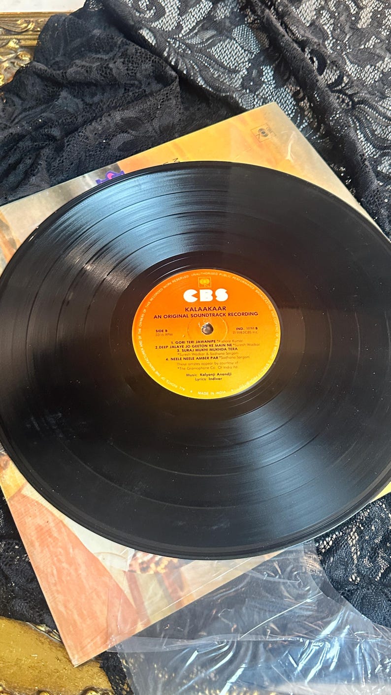 May include: A vintage vinyl record from the soundtrack of "Kalaakaar" by CBS. The black record is centered on a colorful label with text. The record is partially visible, with the album cover and a black lace fabric in the background.