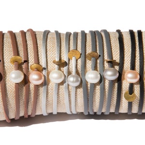 May include: A collection of leather bracelets with gold accents and pearl embellishments. The bracelets are displayed on a fabric-covered form.