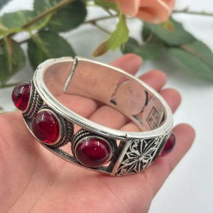 May include: Silver-toned hinged bracelet with six round, deep red gemstones. The bracelet features intricate filigree and decorative patterns. The gemstones are set in a bezel setting. A classic piece of jewelry.