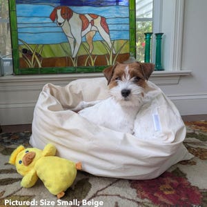 May include: A small, beige dog bed with a white dog inside and a yellow duck toy. The dog bed is round and plush. The dog is a Russell Terrier. The text on the image says "Pictured: Size Small; Beige" and "Reference: 14 lb Russell Terrier."