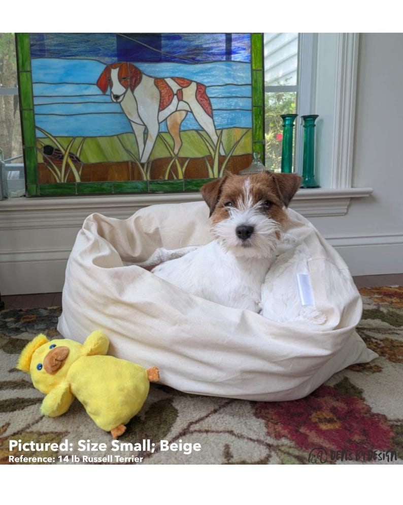 May include: A small, beige dog bed with a white dog inside and a yellow duck toy. The dog bed is round and plush. The dog is a Russell Terrier. The text on the image says "Pictured: Size Small; Beige" and "Reference: 14 lb Russell Terrier."