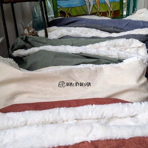 May include: A stack of pet beds in various colors, including blue, black, green, beige, and orange. Each bed features a soft, plush lining. The beds are arranged in a layered display, showcasing the different color options and textures. The text "DENS BY DESIGN" is visible on one of the beds.