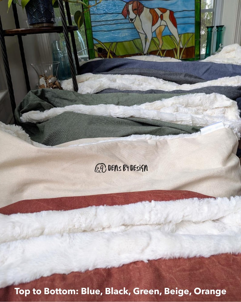 May include: A stack of pet beds in various colors, including blue, black, green, beige, and orange. Each bed features a soft, plush lining. The beds are arranged in a layered display, showcasing the different color options and textures. The text "DENS BY DESIGN" is visible on one of the beds.