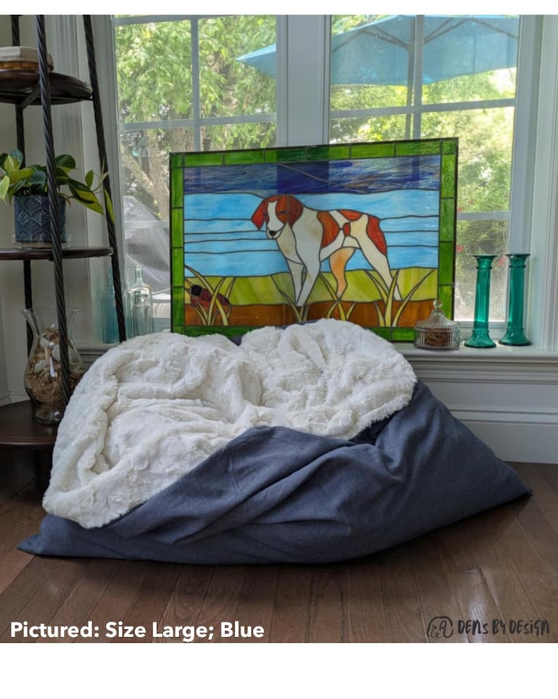 May include: A large blue pet bed with a white, fluffy top. The bed is shown in a room with a stained glass window featuring a dog. The text "Pictured: Size Large; Blue" is at the bottom.