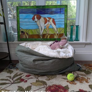 May include: A green dog bed with a white, fluffy interior, a pink stuffed dinosaur, and a tennis ball. A stained glass window with a dog image is in the background. The text "Pictured: Size Medium; Green" is at the bottom.
