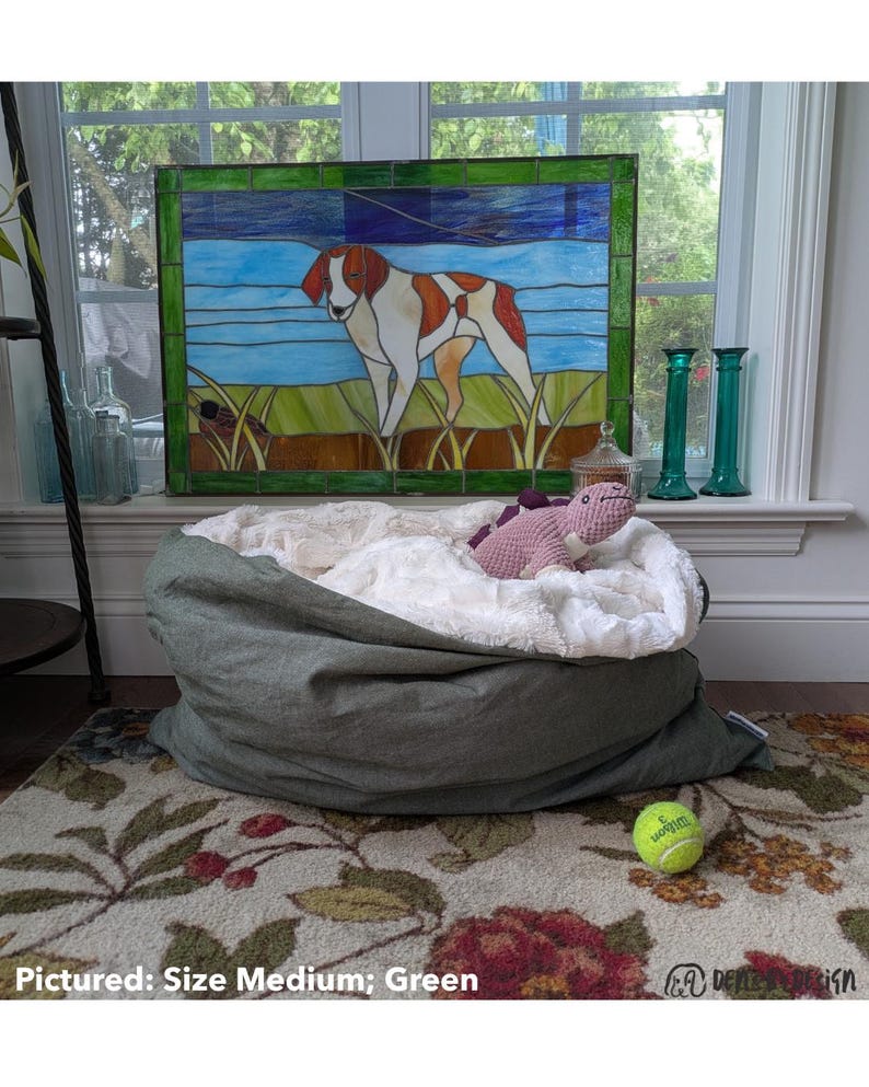 May include: A green dog bed with a white, fluffy interior, a pink stuffed dinosaur, and a tennis ball. A stained glass window with a dog image is in the background. The text "Pictured: Size Medium; Green" is at the bottom.