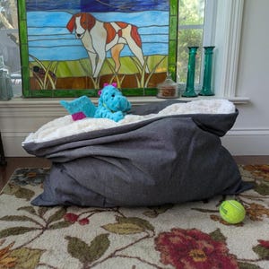 May include: A medium-sized, black pet bed with a plush, white interior. A blue stuffed animal and a pink toy are inside the bed. A yellow tennis ball rests on the floral patterned rug. The text "Pictured: Size Medium; Black" is at the bottom.