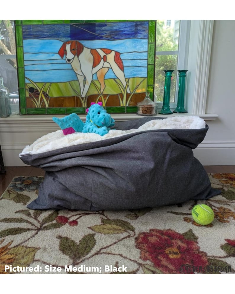 May include: A medium-sized, black pet bed with a plush, white interior. A blue stuffed animal and a pink toy are inside the bed. A yellow tennis ball rests on the floral patterned rug. The text "Pictured: Size Medium; Black" is at the bottom.