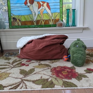 May include: A rust-colored pet bed with a white, fluffy interior sits on a floral rug. A green alligator-shaped pet toy and a small orange and blue ball are nearby. A stained-glass window depicting a dog is in the background.