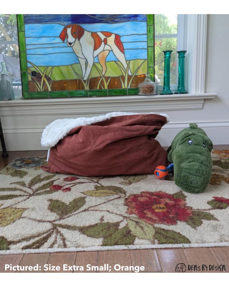 May include: A rust-colored pet bed with a white, fluffy interior sits on a floral rug. A green alligator-shaped pet toy and a small orange and blue ball are nearby. A stained-glass window depicting a dog is in the background.