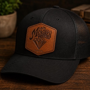 May include: A black baseball cap with a mesh back and a hexagonal tan leather patch on the front. The patch features the text Matt Warren above a diamond graphic and the word diamond below it, all embossed in a dark brown tone on the leather.