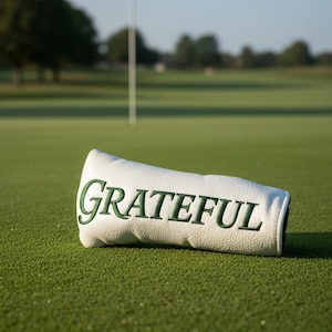 May include: A white golf club head cover with the word "GRATEFUL" embroidered in green. The cover is lying on a green golf course, with a flag and trees in the background.