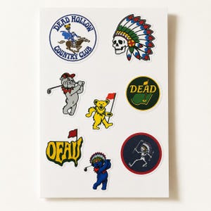 May include: A sticker sheet featuring eight distinct designs including a Dead Hollow Country Club logo, a skull wearing a colorful feathered headdress, a bulldog golfer, a yellow dancing bear with a flag, a Dead golf green, an OFAU logo, and a skeleton.