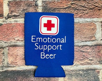 Op de afbeelding: Een blauwe bierblikjeskoeler met de woorden "Emotional Support Beer" in witte tekst. Een rood kruissymbool staat in een witte vierkant bovenaan. De achtergrond is een bakstenen muur.