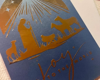 May include: A blue Christmas card with gold foil accents. The card features a nativity scene with a starburst design and the words "Joy to the World". The card is paired with a white envelope.