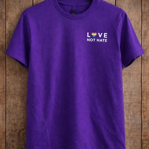Może przedstawiać: Fioletowa koszulka z białym napisem "LOVE NOT HATE". Słowo "LOVE" zawiera tęczowe serce. Koszulka wisi na drewnianym wieszaku na tle drewnianego tła.