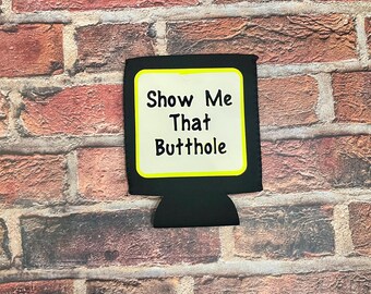 Può includere: Isolante per bevande nero con la scritta "Show Me That Butthole" in bianco con bordo giallo. L'isolante è contro uno sfondo di muro di mattoni.
