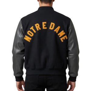 May include: Black varsity jacket with gray leather sleeves. The back of the jacket features the arched text "NOTRE DAME" in gold lettering. The jacket has a black ribbed collar and cuffs.