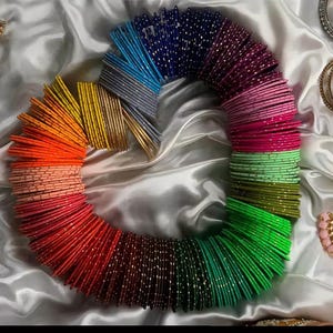 May include: A vibrant collection of colorful bangles arranged in a circular shape. The bangles display a spectrum of colors, including red, orange, yellow, green, blue, and purple, with a shimmering, metallic finish. The bangles are displayed on a white, silky fabric.