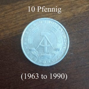 May include: A silver 10 Pfennig coin from the German Democratic Republic, with text reading "10 Pfennig" and "(1963 to 1990)". The coin features a hammer and compass within a wreath, surrounded by the words "DEUTSCHE DEMOKRATISCHE REPUBLIK".
