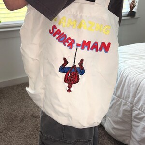 May include: White tote bag with cream-colored straps featuring the text "AMAZING SPIDER-MAN" in yellow and red embroidery. The bag has an embroidered image of Spider-Man hanging upside down.